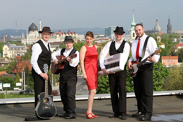 Die DRK Band posiert auf einer Dachterrasse, im Hintergrund ist die Kulisse Dresdens zu sehen. Vier Männer halten ihre Instrumente in den Händen, in ihrer Mitte steht die Sängerin.