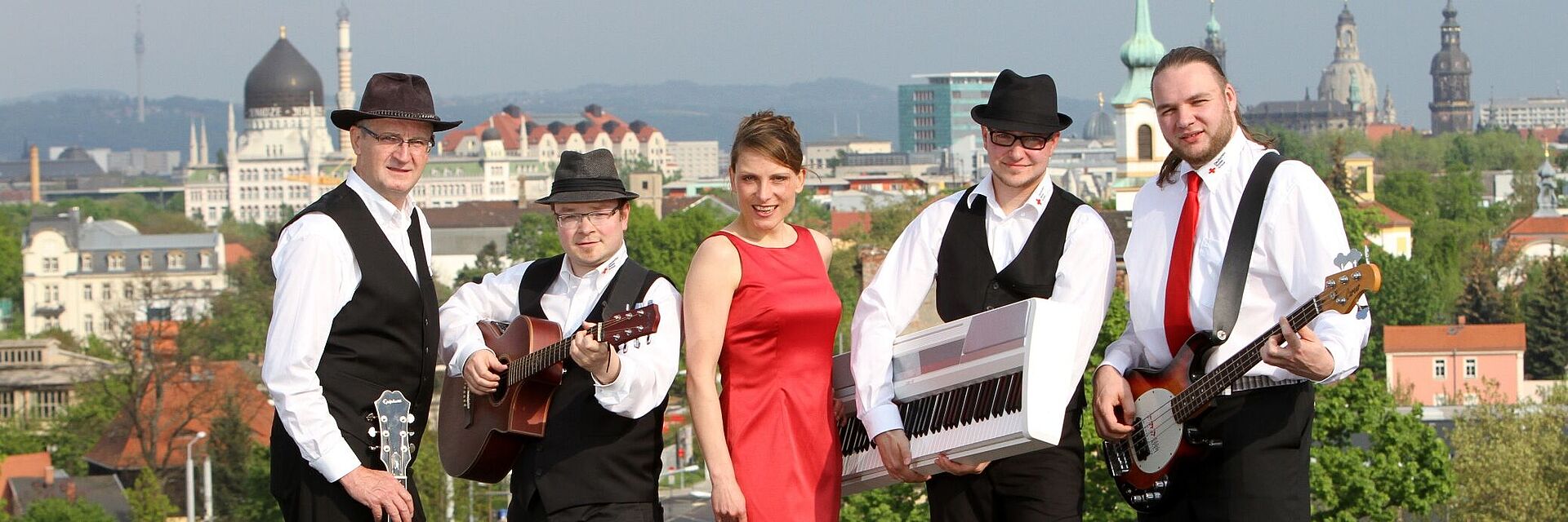 Die DRK Band posiert auf einer Dachterrasse, im Hintergrund ist die Kulisse Dresdens zu sehen. Vier Männer halten ihre Instrumente in den Händen, in ihrer Mitte steht die Sängerin.