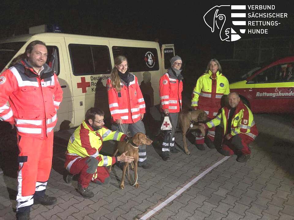 Die Mehrere Personen und ihre Suchhunde der Rettungshundestaffeln Sachsens sind im Einsatz, um vermisste Personen zu finden. Hier stehen sie vor ihrem Einsatzwagen, es ist bereits dunkel.