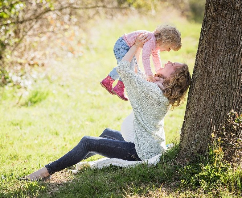 Eine schwangere Frau sitzt an einem Baum und hebt ihr etwa zweijähriges Kind spielerisch hoch. Sie lachen gemeinsam.