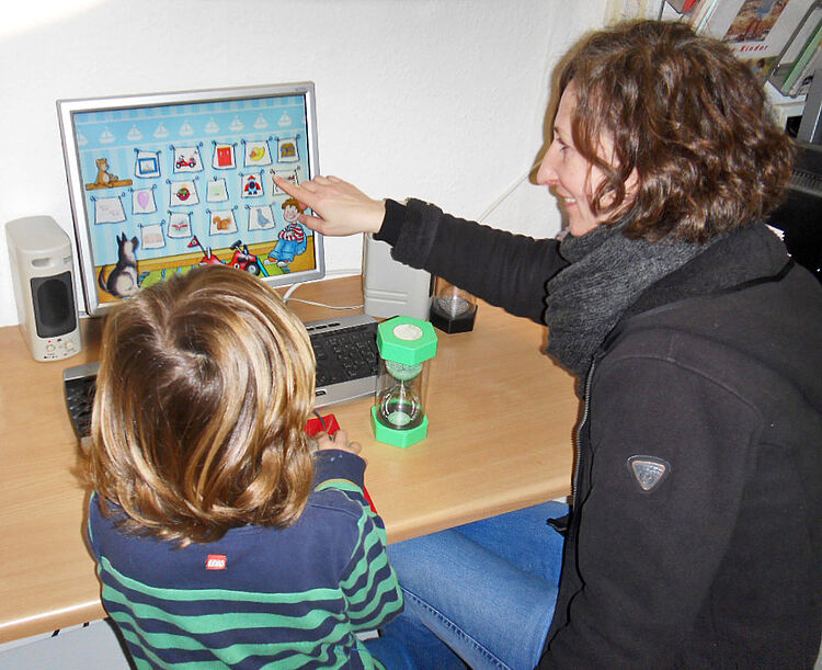 Constanze sitzt mit einem Mädchen am Computer vor einem Lernprogramm. Sie zeigt dem Mädchen Symbole auf dem Bildschirm.