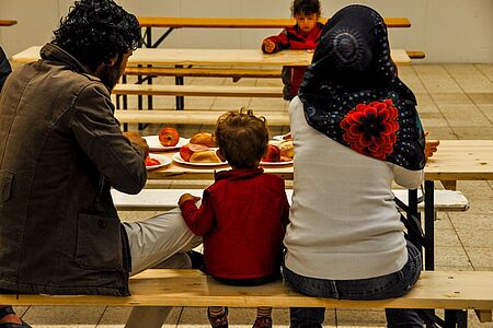 Eine geflüchteter Mann mit seiner Frau und seinem Kind sitzen in einer Erstaufnahmeeinrichtung nebeneinander auf einer Bank und essen Mandarinen.