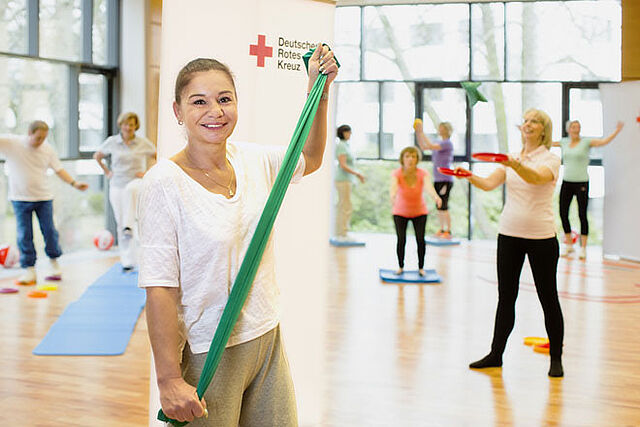 Eine Frau hält ein Fitnessband in den Händen. Im Hintergrund sieht man Teilnehmer eines Kurses auf und neben ihren Fitnessmatten.