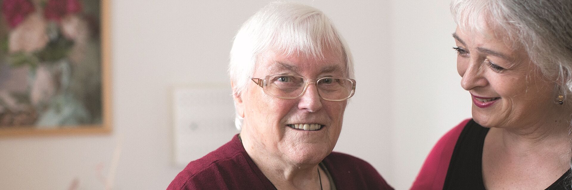 Eine Pflegerin hält den rechten Arm um den Rücken einer Seniorin. Sie wirken vertraut und lächeln.