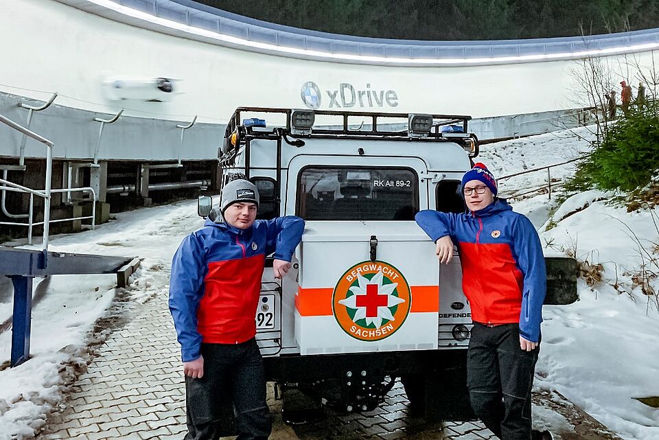 Zwei Kollegen der Bergwacht stehen an ihrem Jeep neben der Eisbahn. Es fährt gerade ein Bob durch die Bahn.