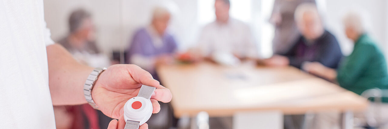 Eine Person hält einen Hausnotruf-Knopf in der Hand. Im Hintergrund sieht man Senioren am Tisch sitzen.