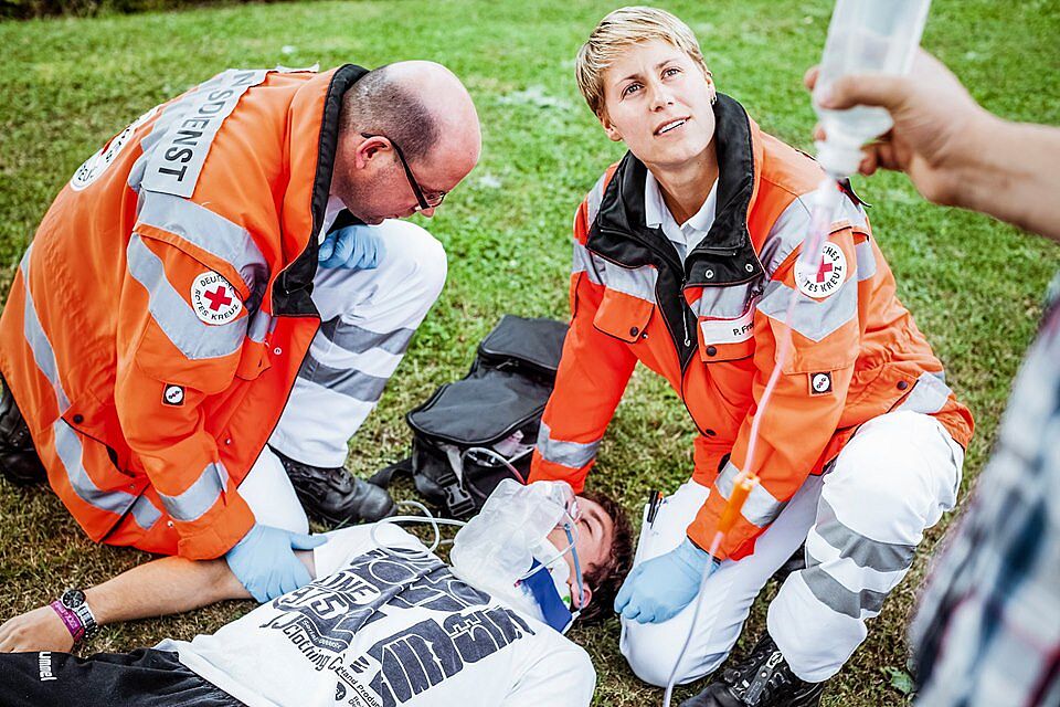 Eine Helferin des Rettungsdienstes versorgt einen jungen Mann, der eine Halskrause trägt. Der junge Mann liegt auf einer Wiese. Auch ein zweiter Helfer ist vor Ort. Eine dritte, nicht erkenntliche Person, hält einen Tropf in der Hand.