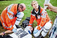 Eine Helferin des Rettungsdienstes versorgt einen jungen Mann, der eine Halskrause trägt. Der junge Mann liegt auf einer Wiese. Auch ein zweiter Helfer ist vor Ort. Eine dritte, nicht erkenntliche Person, hält einen Tropf in der Hand.